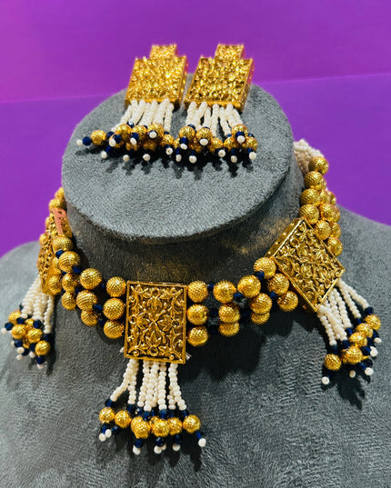 An ornate gold-toned jewelry set displayed on a gray mannequin bust against a purple background. The set includes a beaded necklace with rectangular filigree-style gold pendants, matching drop earrings, and white and navy blue bead tassels.